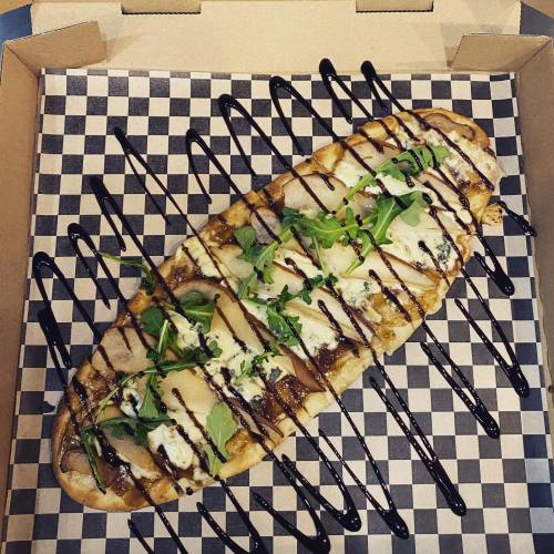 A flatbread topped with cheese, arugula, and drizzled with balsamic glaze, served in a checkered takeout box.