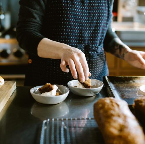 A person in an apron places pieces of baked goods into small bowls on a kitchen counter.
