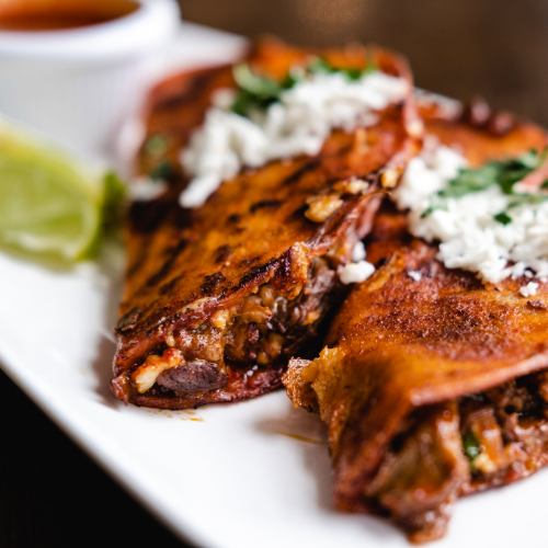 A plate of crispy quesadillas filled with meat, topped with cheese and cilantro, served with a side of sauce and lime.