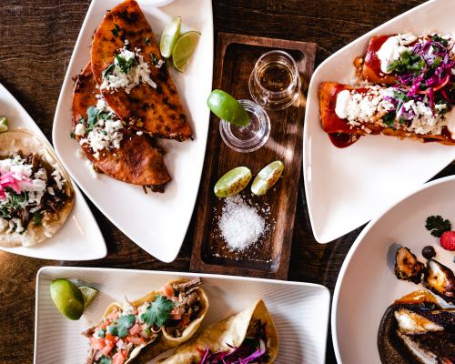 A vibrant spread of tacos, enchiladas, and garnishes, accompanied by lime wedges and a shot of tequila on a wooden tray.