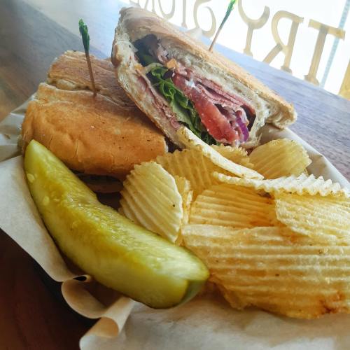 A sandwich with layers of meat and lettuce, served with potato chips and a pickle on the side.