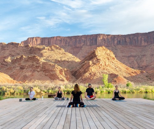 A group of five people practicing yoga on a wooden deck by a river, surrounded by red rock formations and blue skies.