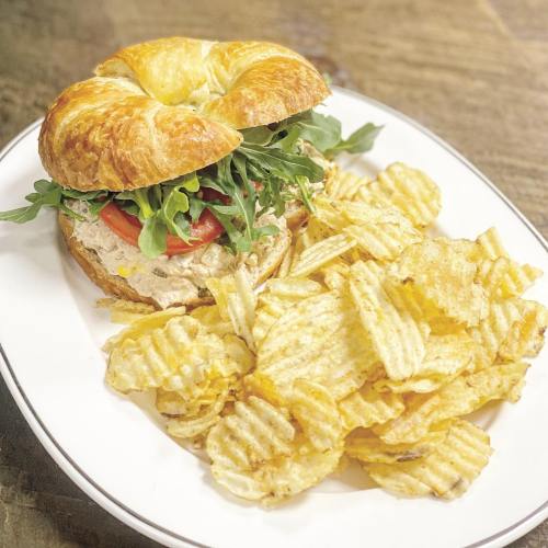 A croissant sandwich filled with tuna, arugula, and tomato, served with a side of wavy potato chips on a white plate.