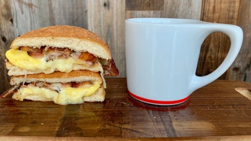 A stacked sandwich with cheese, bacon, and egg next to a white coffee mug on a wooden surface.