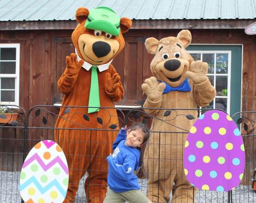 A girl poses playfully with two bear mascots in front of a wooden building, surrounded by colorful Easter eggs.