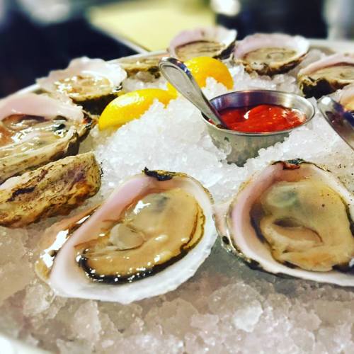 A platter of fresh oysters on ice, garnished with lemon wedges and a small bowl of cocktail sauce.
