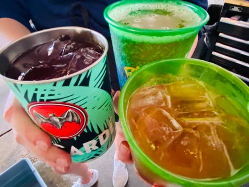 Three colorful cups filled with drinks and ice, held by a person. One cup is green, one is brown, and one is blue.