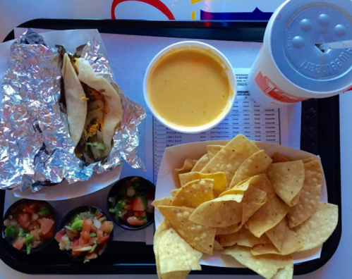 A tray with a taco in foil, cheese soup, tortilla chips, and salsa cups, alongside a drink.