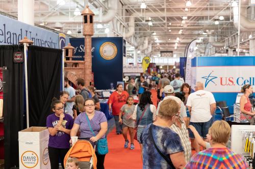 A busy indoor event with people walking through booths and displays, including a castle structure and various banners.