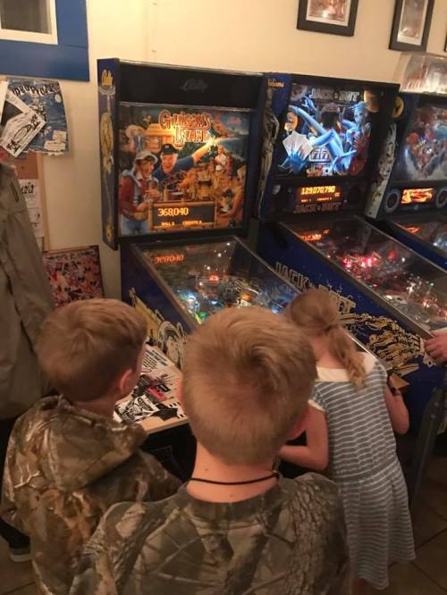 Three children play pinball at an arcade, focused on the game with colorful machines in the background.
