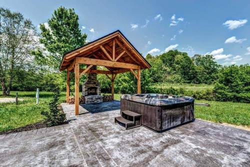 A wooden gazebo with a hot tub and stone fireplace, surrounded by green grass and trees under a clear blue sky.
