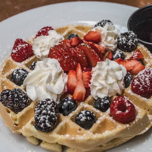 A delicious waffle topped with strawberries, blueberries, blackberries, raspberries, and whipped cream, dusted with powdered sugar.