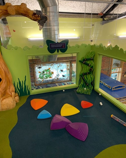 Colorful play area with soft shapes on the floor, green walls, and a large aquarium visible through glass windows.