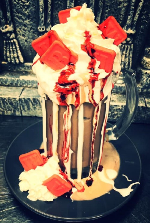 A tall glass filled with chocolate milkshake, topped with whipped cream, red syrup, and red candy pieces, on a black plate.