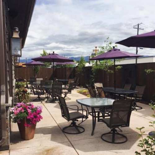 Outdoor patio with tables, chairs, and purple umbrellas, surrounded by greenery and flowers under a cloudy sky.