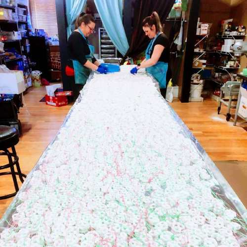 Two women in aprons prepare a large table covered with white and green candy, surrounded by kitchen supplies.