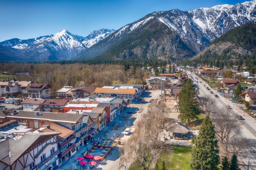 Aerial view of a charming town with colorful buildings, surrounded by mountains and trees under a clear blue sky.