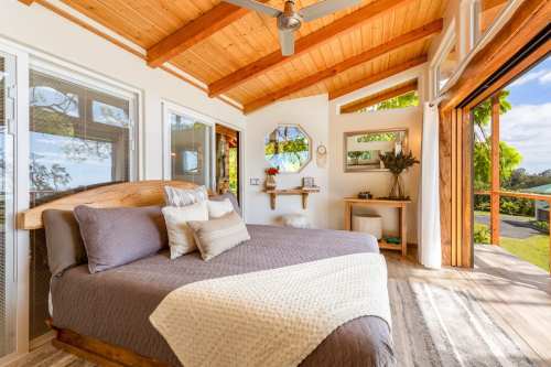 Cozy bedroom with a wooden bed, large windows, and natural light, featuring a minimalist decor and a view of greenery.