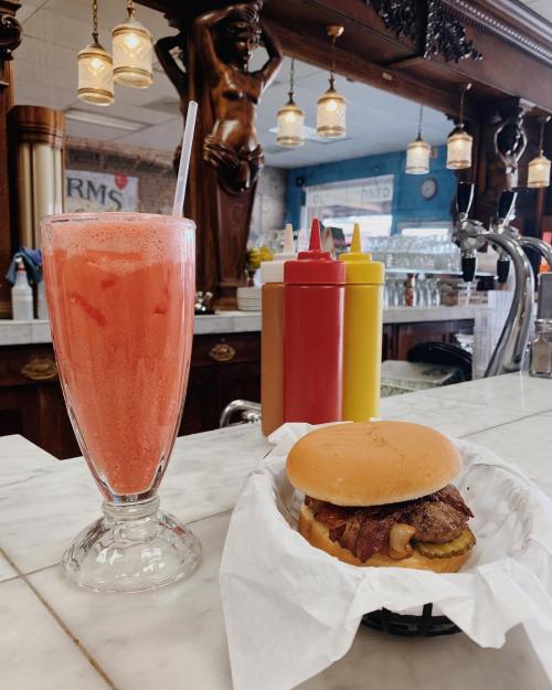 A frosty pink drink and a burger with bacon and pickles on a marble counter in a vintage diner setting.