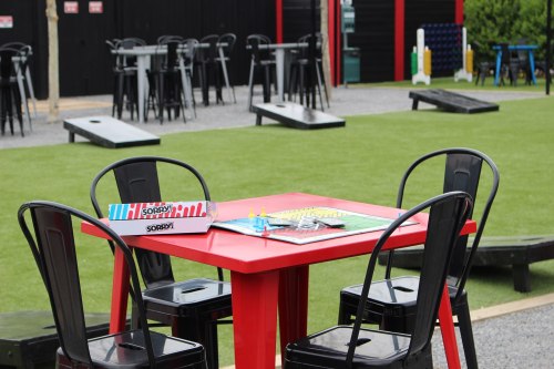 A red table with board games sits in an outdoor area with green grass and black chairs, surrounded by seating and games.