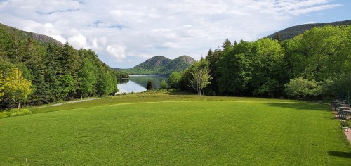 Lush green field with trees and mountains reflected in a calm lake under a partly cloudy sky.