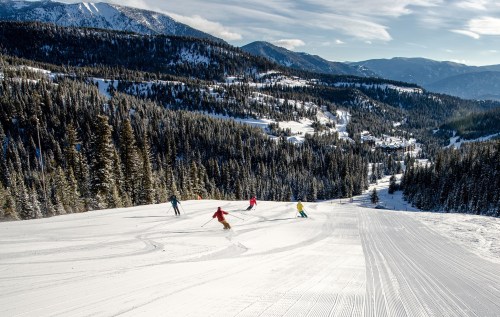 Four skiers navigate a snowy slope surrounded by evergreen trees and mountains under a clear blue sky.