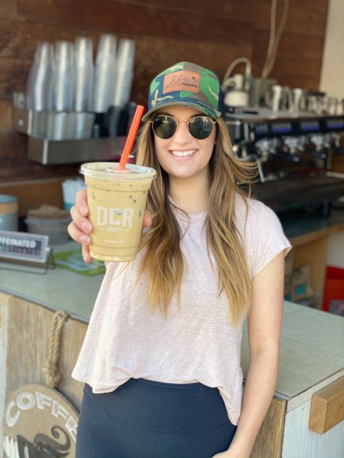 A smiling woman in a cap and sunglasses holds a cup of iced coffee in a café setting.