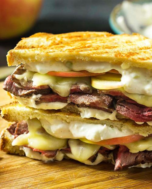 A stacked sandwich with layers of roast beef, cheese, pickles, and creamy sauce on toasted bread, resting on a wooden board.