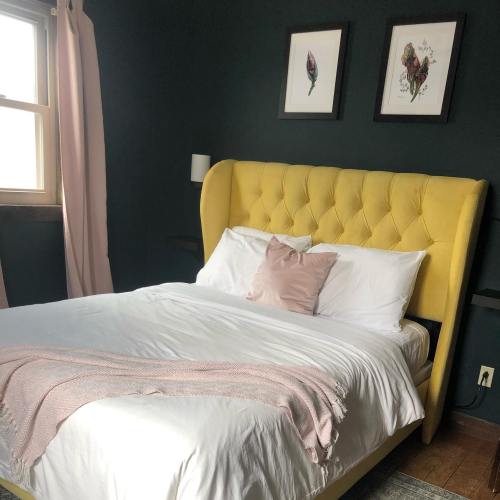 Cozy bedroom with a yellow tufted headboard, white bedding, pink accents, and dark green walls. Natural light from a window.