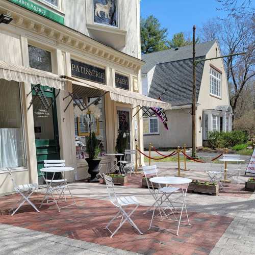 Outdoor seating area with white tables and chairs in front of a patisserie, surrounded by planters and a red rope barrier.