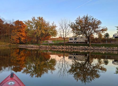 A serene lake scene with autumn trees reflecting in the water, and an RV parked nearby under a clear sky.