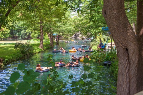 People floating on inner tubes in a lush, green river surrounded by trees and a sunny sky.