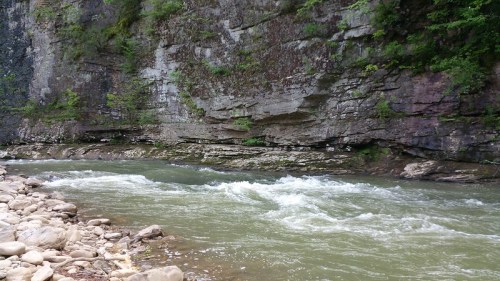 A flowing river surrounded by rocky cliffs and lush greenery, with smooth stones lining the water's edge.