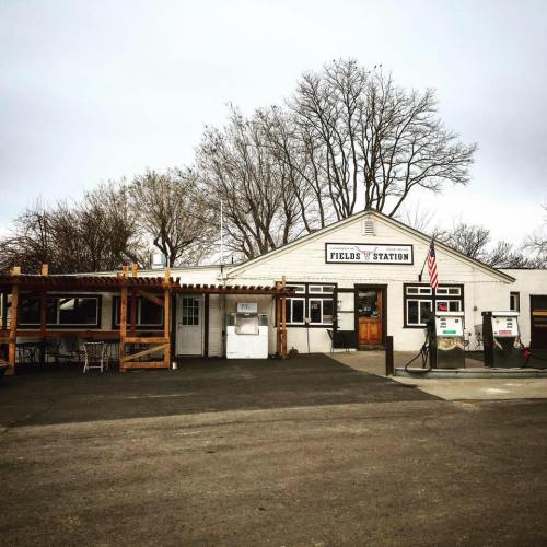 A rustic gas station with a wooden patio, featuring an American flag and surrounded by bare trees under a cloudy sky.