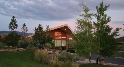 Buffalo Bill Center of the West at dusk, surrounded by greenery and mountains under a cloudy sky.
