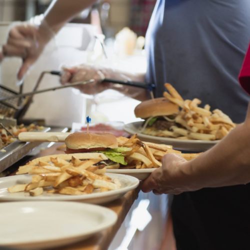 A person holds a plate with a burger and fries, while another prepares food in the background.
