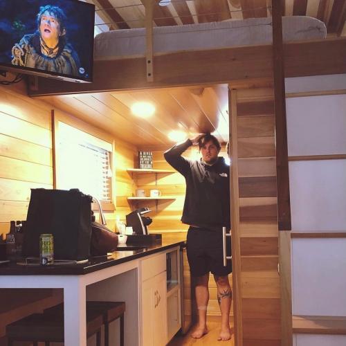 A person stands in a cozy cabin kitchen, looking surprised, with a TV showing a scene from a movie in the background.