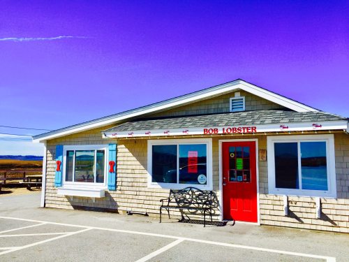 A quaint seafood restaurant named "Bob Lobster" with a red door and blue shutters against a bright purple sky.