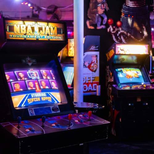A vibrant arcade scene featuring an NBA Jam game cabinet and various classic arcade machines in the background.