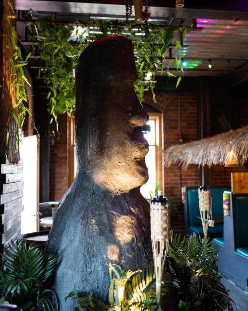 A large stone statue resembling a Moai, surrounded by tropical plants and tiki torches in a vibrant restaurant setting.
