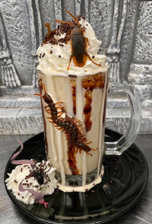 A spooky dessert featuring a tall glass of ice cream topped with whipped cream, chocolate syrup, and creepy crawlers.