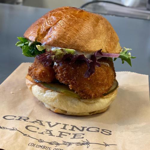 A crispy fried chicken sandwich with lettuce, pickles, and mayo on a toasted bun, served on a paper with "Cravings Cafe."