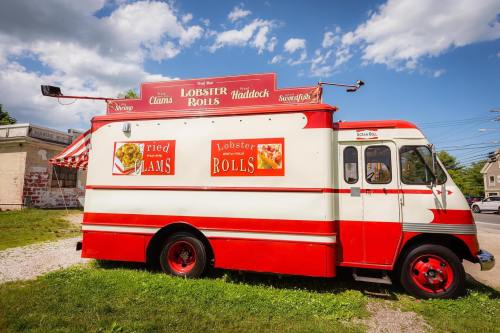 A red and white food truck advertising lobster rolls, fried clams, and haddock, parked on a grassy area under a blue sky.
