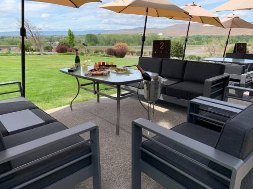 A patio setup with modern seating, a table with snacks, and champagne, surrounded by green grass and umbrellas.