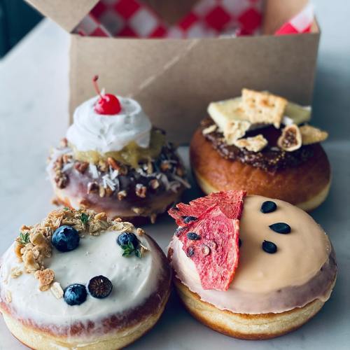 Four colorful donuts with various toppings, including fruit, nuts, and cream, displayed in front of a takeout box.
