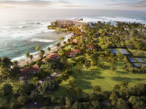 Aerial view of a tropical resort by the ocean, surrounded by palm trees and lush greenery. Waves gently crash on the shore.