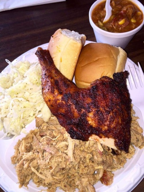 A plate of barbecue chicken, pulled pork, coleslaw, two rolls, and a cup of chili.