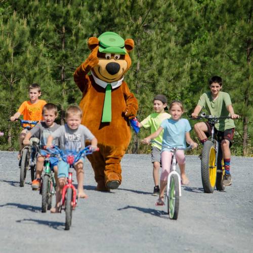 A bear mascot waves as children ride bikes on a gravel path surrounded by trees.