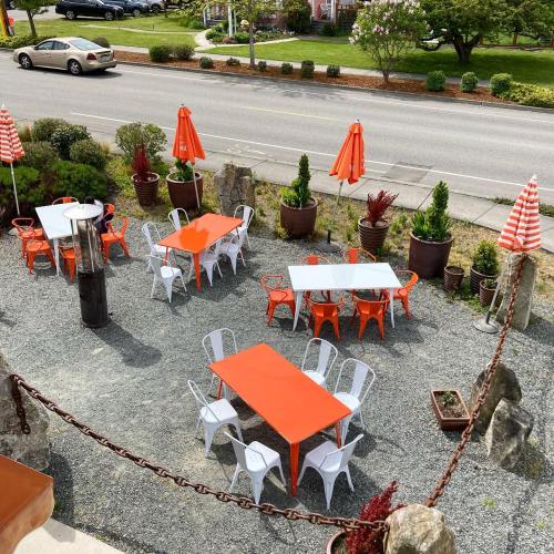 Outdoor dining area with orange and white tables and chairs, surrounded by greenery and umbrellas, near a road.