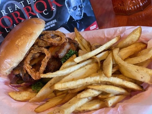 A burger with crispy onion rings and lettuce, served with a side of golden fries, against a horror movie backdrop.